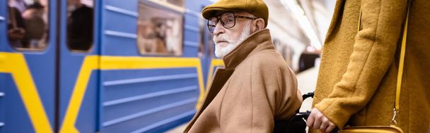 женщина держит инвалидное кресло пожилого мужчины-инвалида на платформе метро возле размытого поезда, баннер - Фото, изображение