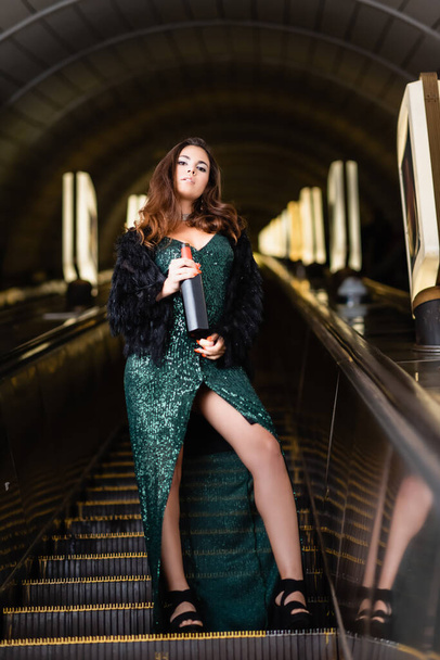 seductive woman in elegant black dress holding bottle of wine on underground escalator - Foto, afbeelding
