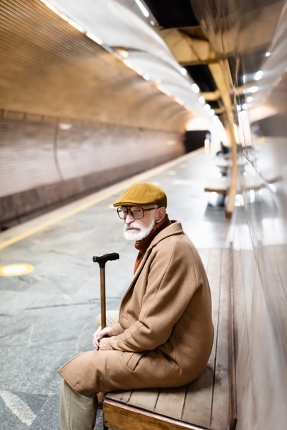 senior man in coat and cap looking away while sitting on underground platform bench - Foto, Imagen