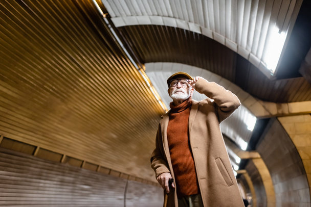 elderly man in autumn clothes touching eyeglasses while standing on metro platform - Valokuva, kuva