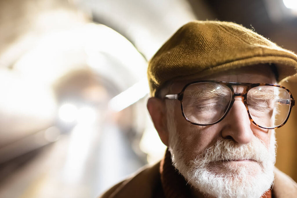 portrait of aged man in cap and eyeglasses standing with closed eyes on metro platform with light in metro tunnel on background - Fotoğraf, Görsel