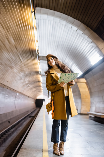 femme élégante en vêtements d'automne regardant loin tout en tenant la carte de la ville sur la plate-forme souterraine - Photo, image