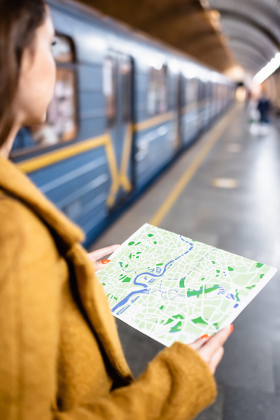 bijgesneden weergave van jonge vrouw met stadsplattegrond terwijl ze in de buurt van metro trein op wazig voorgrond - Foto, afbeelding