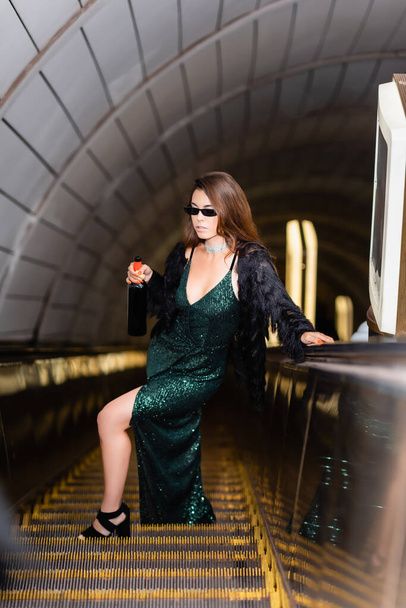 κομψή γυναίκα στις κυλιόμενες σκάλες σε μακρύ μαύρο φόρεμα και γυαλιά ηλίου κρατώντας μπουκάλι κρασί σε θολή πρώτο πλάνο - Φωτογραφία, εικόνα