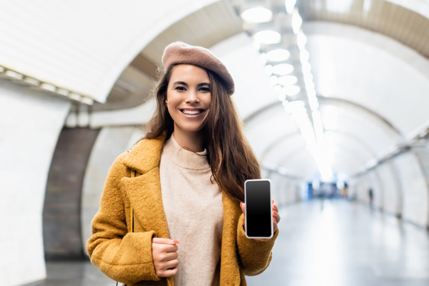 cheerful woman in autumn coat and beret holding smartphone with blank screen at underground station - Foto, Imagen