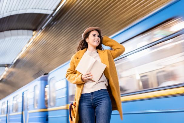 donna felice in abito autunnale tenendo libro e guardando il treno della metropolitana che arriva sulla piattaforma - Foto, immagini