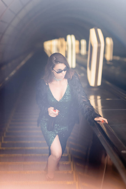 verführerische Frau im schwarzen Kleid, Weinflasche in der Hand und wegschauend auf Rolltreppe, verschwommener Vordergrund - Foto, Bild