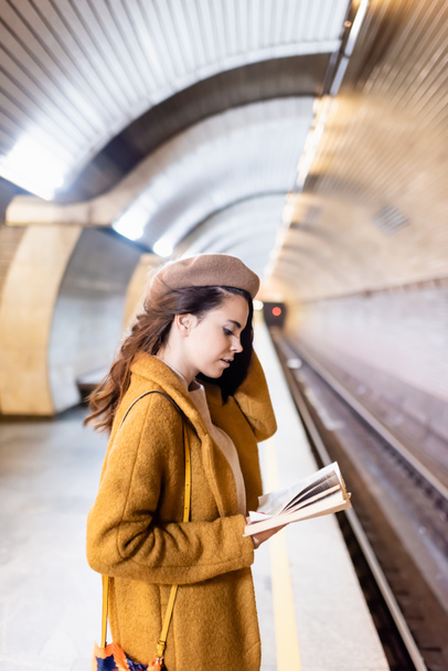 young woman in coat and beret reading book on underground platform - Foto, afbeelding