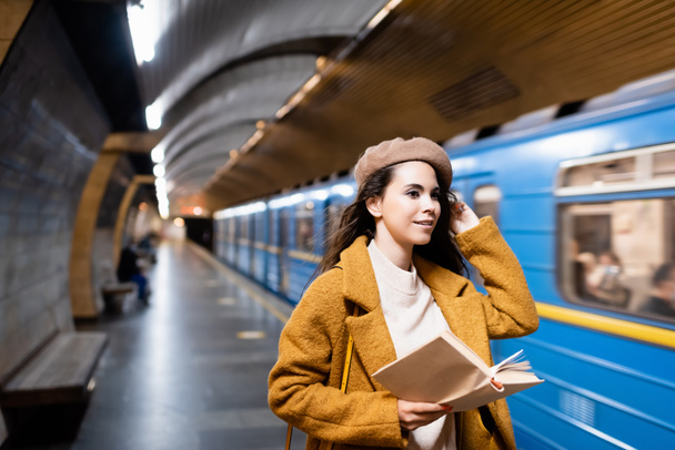 smiling woman in autumn outfit holding book while looking at blurred train on metro platform - Foto, Imagen
