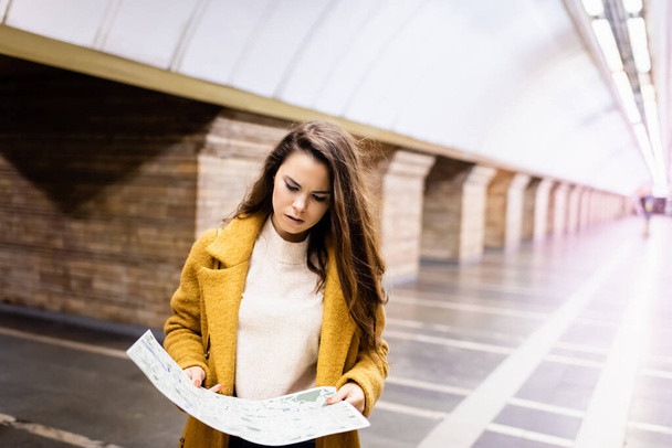 νεαρή γυναίκα σε φθινοπωρινό παλτό σπουδάζει χάρτη της πόλης στο υπόγειο σταθμό - Φωτογραφία, εικόνα