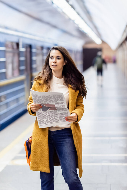 young woman in autumn outfit holding newspaper near blurred metro train on platform - Фото, изображение