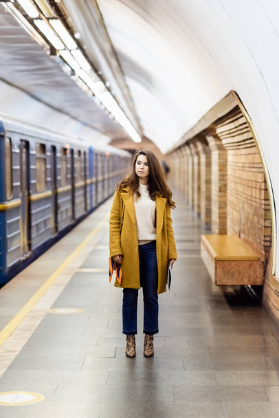 κομψή γυναίκα το φθινόπωρο ρούχα κοιτάζοντας το τρένο στην υπόγεια πλατφόρμα, θολή φόντο - Φωτογραφία, εικόνα
