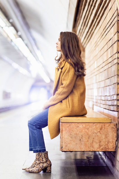 oldalnézetben fiatal nő elegáns őszi ruhák ül földalatti platform padon - Fotó, kép