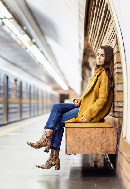 вдумчивая женщина в осеннем наряде сидит на скамейке метро с размытым поездом - Фото, изображение