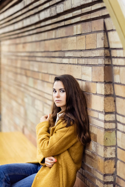 young woman in autumn coat looking at camera while sitting on subway platform bench on blurred background - 写真・画像