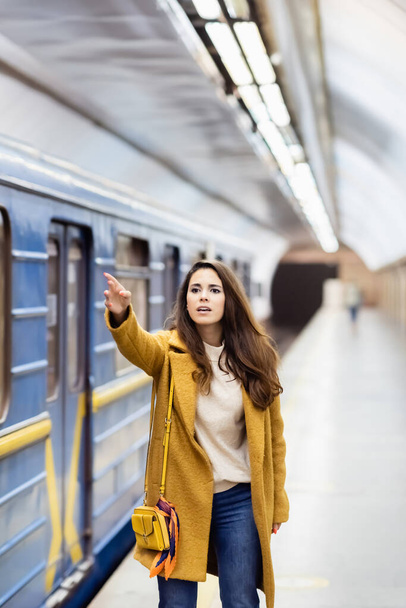 young woman in autumn outfit gesturing with outstretched hand near blurred train on metro platform  - Фото, изображение