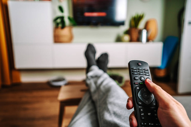 Un homme se détendre sur le canapé et utiliser la télécommande et regarder la télévision dans une maison avec un intérieur confortable et accueillant - Photo, image