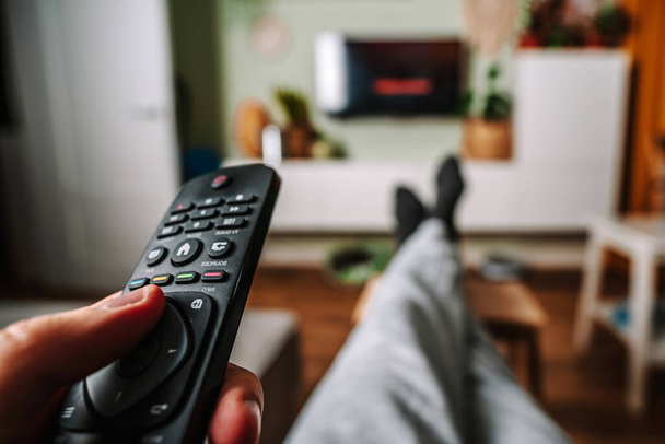 Un hombre se está relajando en el sofá y utilizar el control remoto y ver la televisión en una casa con un acogedor interior acogedor - Foto, imagen