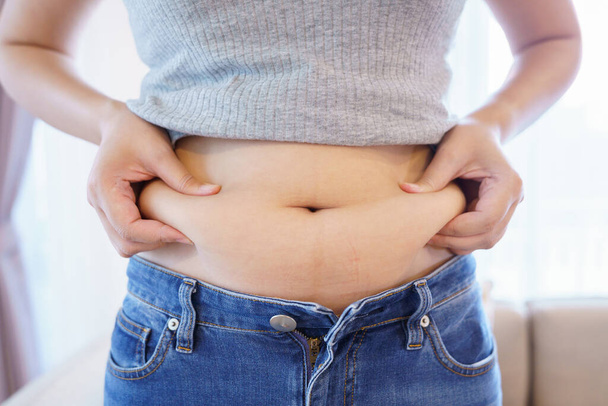 女性の体脂肪腹。お腹の脂が多すぎる肥満の女性の手。お腹を減らし健康な胃の筋肉を形成するための食生活の概念. - 写真・画像