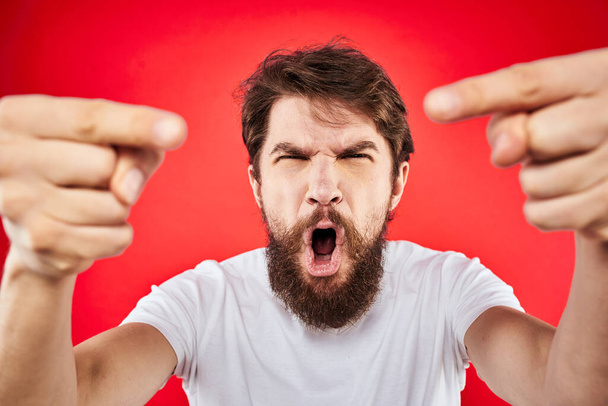 szakállas férfi gesztikulál kezével fehér póló érzelmek arc kifejezés agresszió piros elszigetelt háttér - Fotó, kép