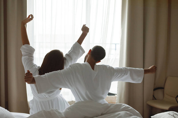 Ζευγάρι άντρας και γυναίκα με λευκά παλτά που κάθονται στο κρεβάτι αφού ξυπνήσουν και τεντωθούν μπροστά από το παράθυρο στο δωμάτιο του ξενοδοχείου. Πίσω όψη. - Φωτογραφία, εικόνα