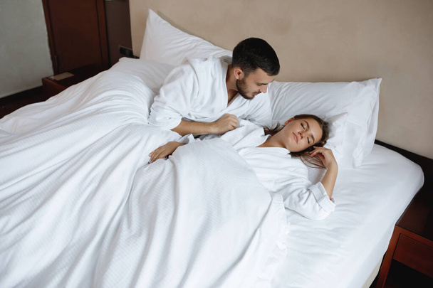 Pareja en la cama. Joven barbudo hombre y mujer yacen en una almohada blanca y se despiertan por la mañana temprano. - Foto, Imagen