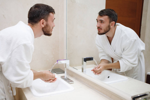 Бородатый парень в белом халате моется в раковине с краном в ванной. - Фото, изображение