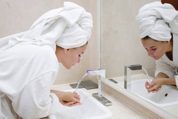 Mujer joven con una bata blanca se lava en el lavabo con un grifo en el baño. Reflexión en el espejo. Enfoque seleccionado. - Foto, Imagen