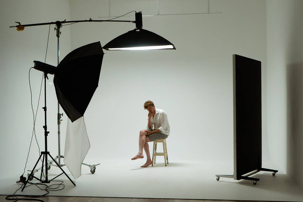 Young barefoot man sits on chair in pose of thinker posing in photo studio. Male model wearing white shirt on white background among studio equipment and lighting fixtures. Backstage concept - 写真・画像