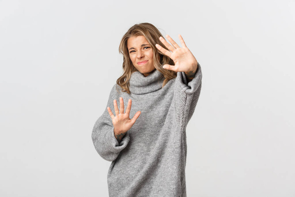 Image of disturbed blond woman feeling uncomfortable and asking to stop photographing, stretching hands forward to cover face from light, standing over white background - Photo, Image