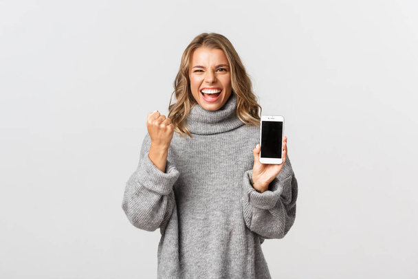 灰色のセーターで幸せな女性を獲得し、達成に勝利し、スマートフォンの画面を示し、拳ポンプを作り、白い背景 - 写真・画像