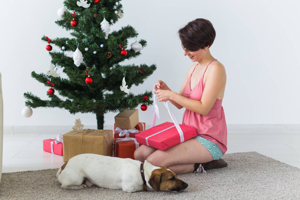 Szczęśliwa kobieta z psem otwierającym prezenty świąteczne. Choinka z prezentami pod spodem. Dekorowany salon - Zdjęcie, obraz