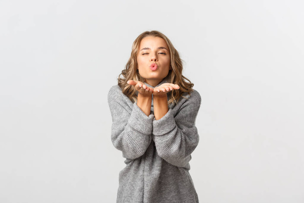 Portrait of attractive lovely woman in grey sweater sending air kiss, standing over white background - 写真・画像