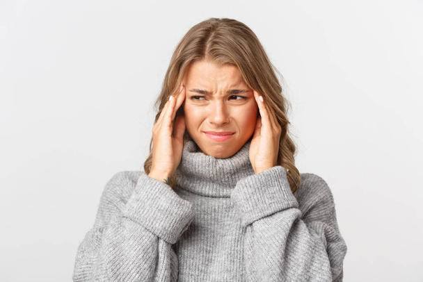 Close-up of grimacing young woman in grey sweater, grimacing and holding hands on head, having painful headache, standing over white background - Photo, Image