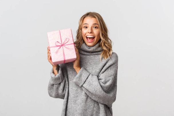 Retrato de chica rubia atractiva en suéter gris, mirando feliz, recibiendo regalo de cumpleaños en caja rosa, de pie sobre fondo blanco - Foto, Imagen
