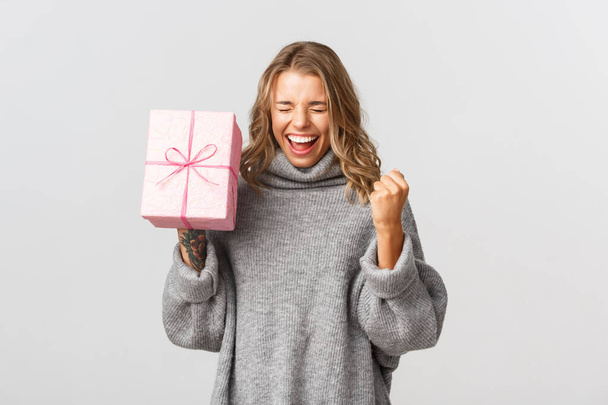 Vidám csinos lány szürke pulóverben, boldognak látszik, ünnepli a születésnapját és ajándékot kap, fehér háttér felett áll - Fotó, kép
