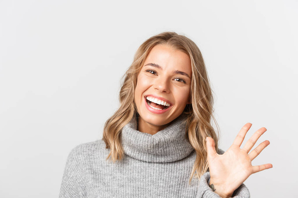 Close-up of friendly attractive girl in grey sweater, smiling and saying hi, waving hand to greet someone, standing over white background - Photo, Image
