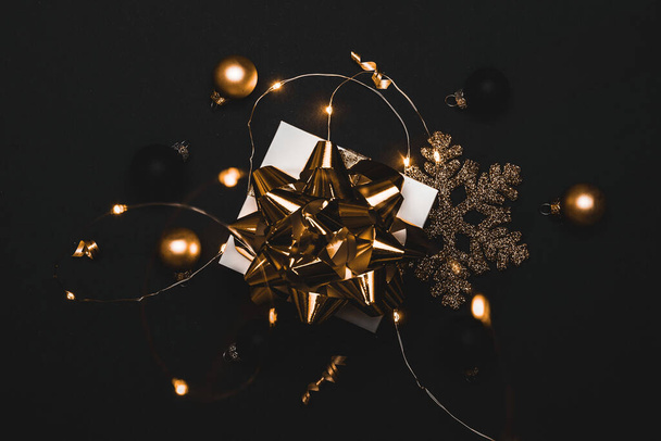 Winterdesign. Weiße Geschenkschachtel mit goldenem Band, Neujahrskugeln und funkelnden Lichtergirlanden in weihnachtlicher Komposition auf schwarzem Hintergrund als Grußkarte. Weihnachtskulisse mit Platz für Text - Foto, Bild