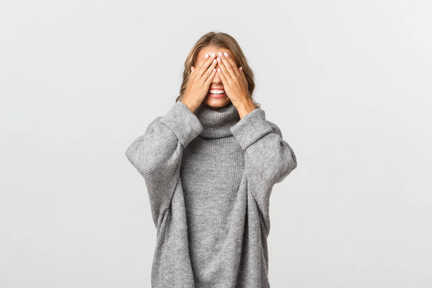 Portrait of beautiful young woman in grey sweater, standing blindsided with hands on face, waiting for surprise, standing over white background - Photo, Image
