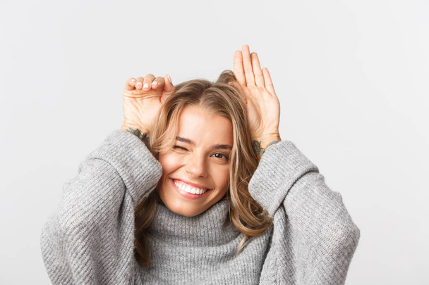 Close-up of lovely blond girl in grey sweater, smiling and showing puppy ears with hands raised over head, standing over white background - Foto, Bild