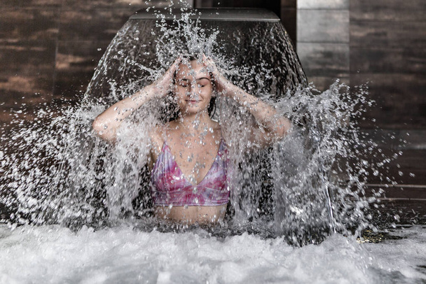Pozitivní mladá samice si užívá procedury vodoléčby a cákání vody, když stojí pod vodopádem v lázeňském bazénu - Fotografie, Obrázek