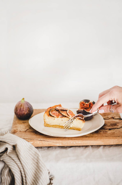 Hand of woman eating homemade gluten free fig cheesecake with fresh figs on white plate over rustic wooden board, white wall at background, copy space. Healthy dessert, artisan sweet food concept - Фото, изображение