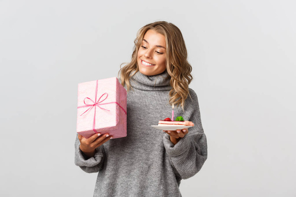 Porträt eines attraktiven blonden Mädchens, das seinen Geburtstag feiert, Geschenk und Kuchen hält und vor weißem Hintergrund steht - Foto, Bild