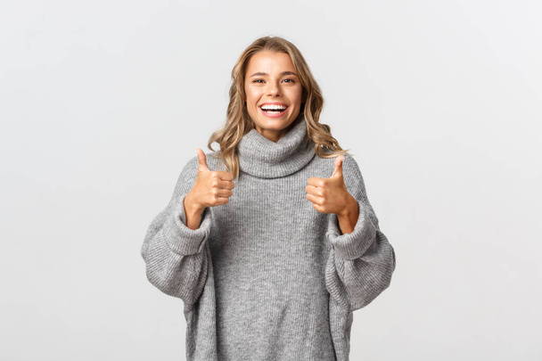 Εικόνα της χαρούμενης ικανοποιημένης γυναίκας με γκρι πουλόβερ, που δείχνει τους αντίχειρες και χαμογελά, συστήνει κάτι, όπως και να εγκρίνει το προϊόν, στέκεται πάνω από το λευκό φόντο - Φωτογραφία, εικόνα