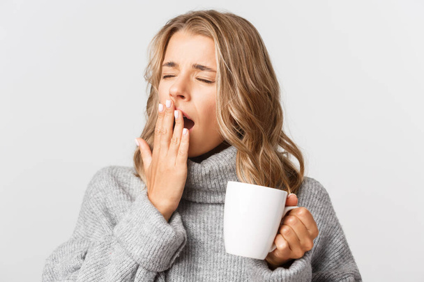 Primer plano de la soñolienta rubia en suéter gris, bebiendo café y bostezando, de pie sobre fondo blanco - Foto, Imagen
