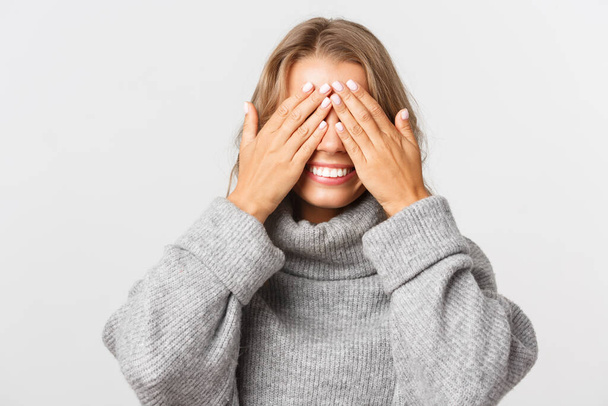 Close-up of happy beautiful woman in grey sweater, standing blindsided with hands on face, waiting for surprise and smiling, white background - Zdjęcie, obraz