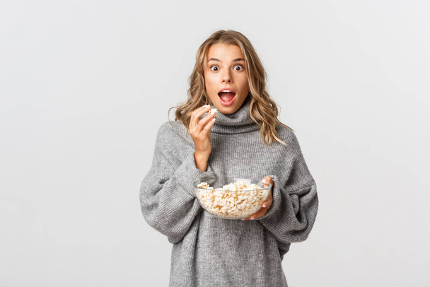 Image of amazed blond girl watching movie, looking excited, eating popcorn, standing over white background - Photo, Image