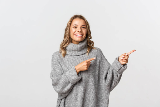 Imagen de una hermosa chica rubia en suéter gris apuntando con los dedos hacia la derecha, mostrando su logotipo o pancarta, de pie sobre fondo blanco - Foto, Imagen