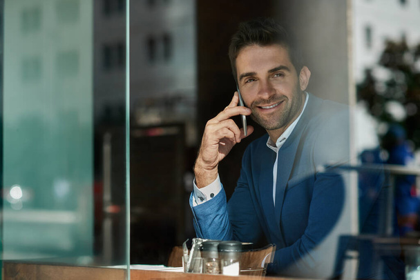 Smiling young businessman talking on his cellphone while sitting by a window inside of a cafe - Fotografie, Obrázek