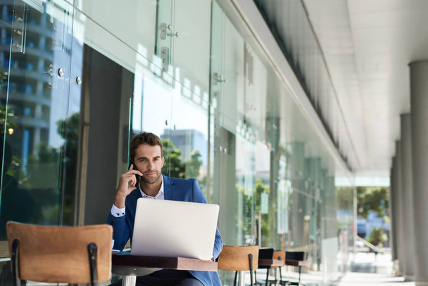Muotokuva nuoresta liikemiehestä, joka puhuu kännykkäänsä ja työskentelee kannettavan kanssa istuessaan ulkona kahvilapöydässä. - Valokuva, kuva
